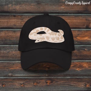 May include: Black baseball cap with a beige and brown snake design. The cap has a curved peak and a classic six-panel construction. The text "Creepy Crawly Apparel" is printed on the top right of the cap.