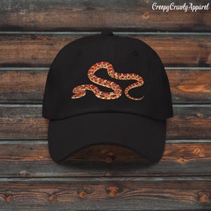 May include: A black baseball cap featuring a graphic print of a coiled snake with orange, red, and tan patterned scales. The text CreepyCrawlyApparel is printed in white in the upper right corner of the image above the cap.
