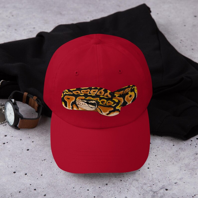 May include: A red baseball cap featuring an embroidered image of a ball python. The snake is depicted in shades of yellow, brown, and black. The cap has a curved brim and a classic six-panel design.