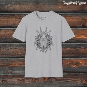 Op de afbeelding: Een grijs T-shirt met een zwart-witte illustratie van een kever omringd door bladeren. De tekst "Creepy Crawly Apparel" is gedrukt in de rechterbovenhoek van de afbeelding.