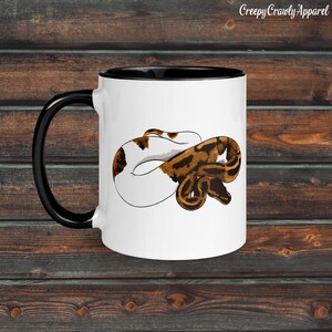 Puede incluir: Taza de cerámica blanca con asa e interior negros. La taza presenta una ilustración detallada de una serpiente marrón y blanca. El texto "Creepy Crawly Apparel" está impreso en la parte superior derecha de la taza.