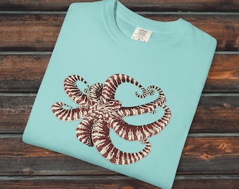 May include: A light blue t-shirt featuring a detailed illustration of an octopus in shades of brown and cream. The shirt has a crew neck and the text "Creepy Crawly Apparel" is printed at the top right.