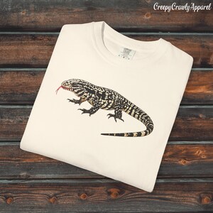 Puede incluir: Camiseta color crema con una ilustración detallada de un lagarto blanco y negro con la lengua roja extendida. La camiseta tiene cuello redondo y el texto "Creepy Crawly Apparel" es visible en la esquina superior derecha.