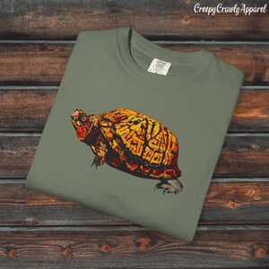 May include: An olive green t-shirt featuring a detailed illustration of a turtle with a yellow and orange shell. The turtle's head and legs are visible, and the shirt has the text "Creepy Crawly Apparel" in the upper right corner.