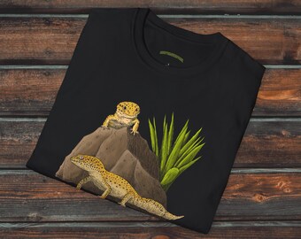 Może przedstawiać: Czarny T-shirt z grafiką przedstawiającą dwa żółto-brązowe gekony na skale z zieloną rośliną. Gekony są zwrócone w różne strony. Koszulka jest złożona na pół i leży na drewnianej powierzchni.