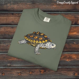 Puede incluir: Camiseta verde oliva con una ilustración detallada de una tortuga. La tortuga tiene un caparazón estampado en marrón y amarillo, y un cuerpo y patas moteados en blanco y gris. El texto "Creepy Crawly Apparel" es visible en la esquina superior derecha.