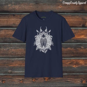 Op de afbeelding: Een donkerblauw t-shirt met een witte afbeelding van een kever omringd door bladeren. De tekst "Creepy Crawly Apparel" is zichtbaar in de rechterbovenhoek van de afbeelding.