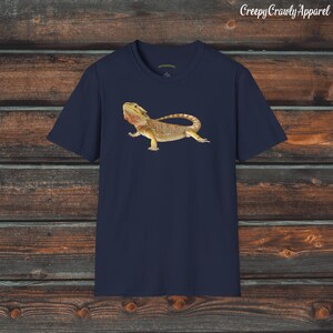 Op de afbeelding: Een donkerblauw t-shirt met een afbeelding van een baardagame. De hagedis is bruin, geel en oranje met een stekelige kam op zijn kop.