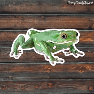 May include: A green frog sticker with white borders, set against a wood grain background. The frog has large, detailed eyes and is in a crawling position. The text "Creepy Crawly Apparel" is in the upper right corner.