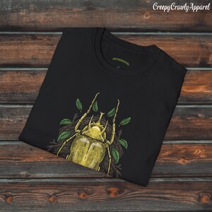May include: Black t-shirt with a yellow beetle graphic. The beetle is surrounded by green leaves and is in a natural setting. The t-shirt is folded and displayed on a wooden surface.