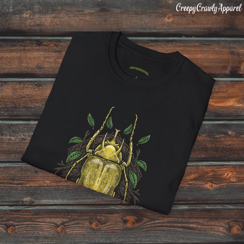 May include: Black t-shirt with a yellow beetle graphic. The beetle is surrounded by green leaves and is in a natural setting. The t-shirt is folded and displayed on a wooden surface.