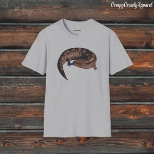 May include: A grey t-shirt with a brown and black lizard graphic. The lizard is curled up with its tail wrapped around its body. The text "Creepy Crawly Apparel" is printed in the top right corner of the image.
