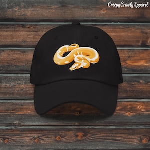 Può includere: Cappellino da baseball nero con un'illustrazione dettagliata di un serpente giallo e bianco. Il serpente è arrotolato e presenta motivi intricati. Il testo "Creepy Crawly Apparel" è stampato in alto a destra sul cappellino.
