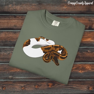 Puede incluir: Una camiseta verde oliva con una ilustración detallada de una serpiente en tonos marrón y blanco. La serpiente está enrollada, con la cabeza y la cola superpuestas. El texto "Creepy Crawly Apparel" está en la esquina superior derecha.
