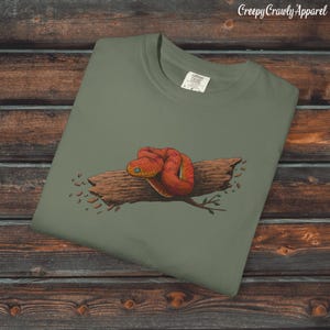 Puede incluir: Una camiseta verde oliva con una ilustración detallada de una serpiente roja enroscada sobre un tronco marrón. La serpiente tiene ojos amarillos y está rodeada de pequeñas hojas marrones. El texto "Creepy Crawly Apparel" está en la esquina superior derecha.