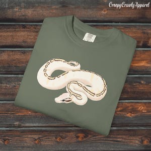 May include: An olive green t-shirt with a cream-coloured snake graphic and brown markings. The snake is coiled on the shirt. The text "Creepy Crawly Apparel" is in the upper right corner.