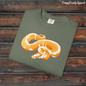 May include: Olive green t-shirt featuring a detailed illustration of an orange and white snake. The snake is coiled and has a detailed pattern. The text "Creepy Crawly Apparel" is in the upper right corner.
