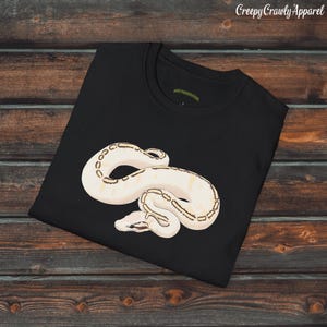 Peut inclure: T-shirt noir avec une illustration d'un serpent crème et des marques marron. Le serpent est enroulé sur le devant. Le texte "Creepy Crawly Apparel" est dans le coin supérieur droit. Le t-shirt est sur une surface en bois.