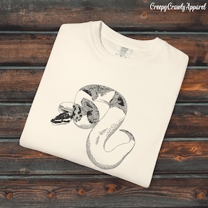 Könnte beinhalten: Cremefarbenes T-Shirt mit einer detaillierten Schwarz-Weiß-Illustration einer zusammengerollten Schlange. Der Kopf der Schlange ist sichtbar, mit komplizierten Mustern auf ihrem Körper. Der Text "Creepy Crawly Apparel" befindet sich oben rechts.