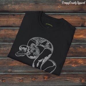 Op de afbeelding: Zwart T-shirt met een witte lijntekening van een slang op de voorkant. De slang is opgerold en heeft een gedetailleerd patroon.