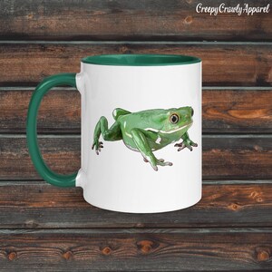 May include: White ceramic mug with a green handle and rim, featuring a detailed illustration of a green frog. The frog has white markings and is set against a white background. The text "Creepy Crawly Apparel" is printed on the top right.