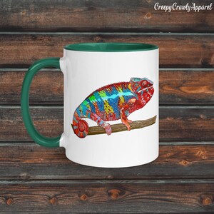 Peut inclure: Mug en céramique blanche avec une anse et un intérieur verts. La tasse présente une illustration colorée d'un caméléon perché sur une branche. Le caméléon a des marques rouges, bleues, vertes et jaunes. Le texte "Creepy Crawly Apparel" est en haut.
