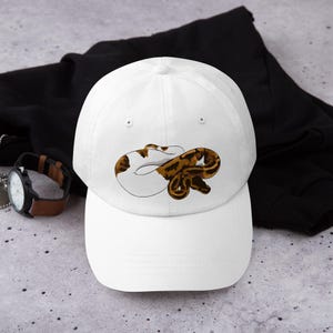 May include: White baseball cap featuring a brown and white snake illustration. The cap has a curved brim and a classic six-panel design. The snake design is detailed with brown and white markings, creating a realistic appearance. This cap is a stylish accessory.