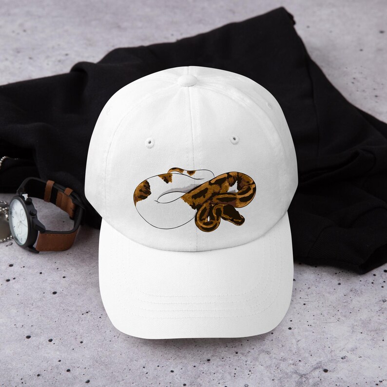 May include: White baseball cap featuring a brown and white snake illustration. The cap has a curved brim and a classic six-panel design. The snake design is detailed with brown and white markings, creating a realistic appearance. This cap is a stylish accessory.