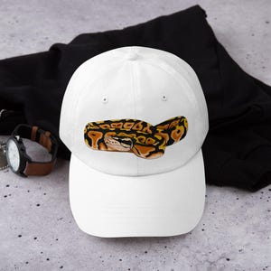 May include: White baseball cap featuring a detailed illustration of a ball python. The snake is depicted with a pattern of black, brown, and yellow markings. The cap is a classic six-panel design.