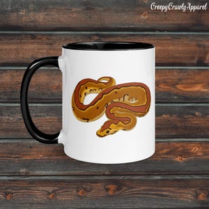 May include: White ceramic mug with a black handle and rim. The mug features a detailed illustration of a brown and orange snake. The text "Creepy Crawly Apparel" is printed on the top right of the mug.