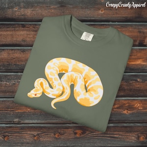 May include: Olive green t-shirt featuring a large, stylized yellow and white snake design. The snake is coiled and detailed, with a red eye. The text "Creepy Crawly Apparel" is printed in the upper right corner.