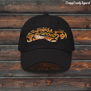 Puede incluir: Gorra de béisbol negra con un diseño de serpiente bordada en tonos amarillos, naranjas y marrones. La gorra tiene una visera curva y una construcción clásica de seis paneles. El texto "Creepy Crawly Apparel" está impreso en la parte superior derecha.