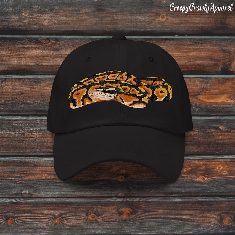 May include: Black baseball cap featuring an embroidered snake design in shades of yellow, orange, and brown. The cap has a curved brim and a classic six-panel construction. The text "Creepy Crawly Apparel" is printed on the top right.