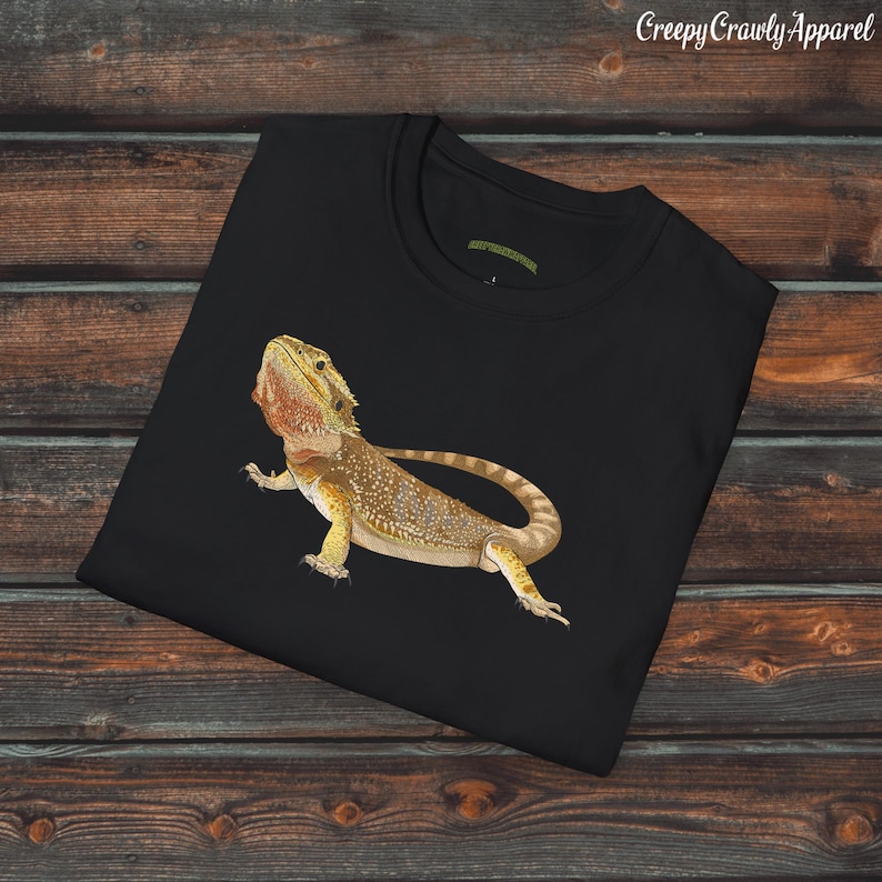 Op de afbeelding: Zwart T-shirt met een afbeelding van een baardagame hagedis. De hagedis is geel en bruin met een stekelige kam op zijn kop. De hagedis kijkt naar de rechterkant van de afbeelding.