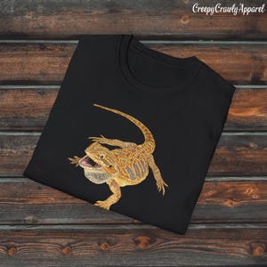 May include: Black t-shirt featuring a graphic of a bearded dragon lizard with a yellow and brown pattern. The lizard is in a defensive posture with its mouth open.
