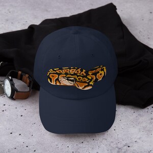 May include: Navy blue baseball cap featuring an embroidered image of a ball python. The snake is depicted in shades of brown, yellow, and black, with detailed scales and markings. The cap is a classic six-panel design with a curved brim.