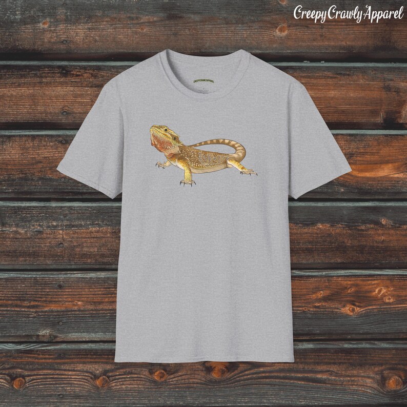 Op de afbeelding: Een grijs t-shirt met een afbeelding van een baardagame. De hagedis is bruin en geel met een stekelige kam op zijn kop. Het t-shirt heeft een ronde hals en korte mouwen.