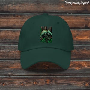 May include: A dark green baseball cap with a detailed illustration of a chameleon in a natural setting. The cap has a curved peak and a classic six-panel design. The text "Creepy Crawly Apparel" is printed on the top right.