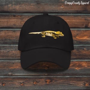 May include: A black baseball cap featuring a centred graphic illustration of a yellow and brown crested gecko. The text CreepyCrawlyApparel is printed in white in the upper right corner of the image above the hat.