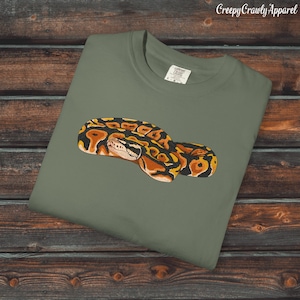 Könnte beinhalten: Ein olivgrünes T-Shirt mit einer detaillierten Illustration einer Königspython. Die Schlange ist mit braunen, schwarzen und gelben Markierungen dargestellt. Das T-Shirt hat einen Rundhalsausschnitt und kurze Ärmel, ideal für den Alltag.