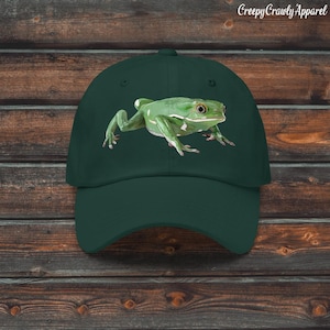 May include: A dark green baseball cap with a detailed illustration of a green frog with white markings. The frog is on the front of the cap. The text "Creepy Crawly Apparel" is printed in white on the top right.