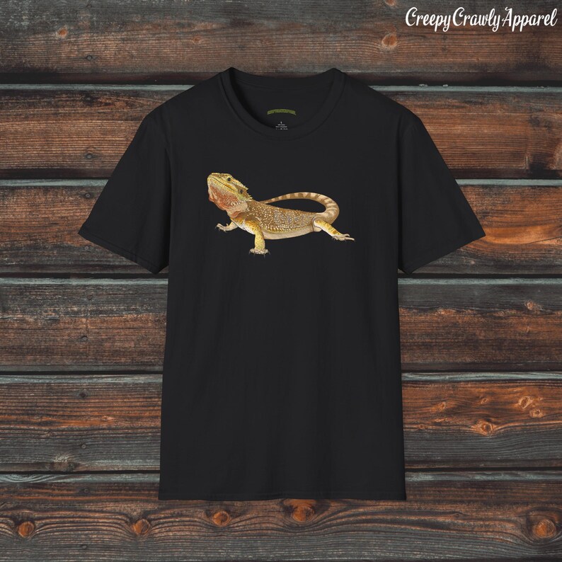 Op de afbeelding: Zwart T-shirt met een afbeelding van een baardagame hagedis. De hagedis is bruin en geel met een stekelige kam op zijn kop. De tekst "Creepy Crawly Apparel" staat in de rechterbovenhoek van de afbeelding.