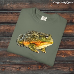 May include: An olive green t-shirt with a detailed illustration of a frog. The frog has a textured, green and yellow body with orange accents. The text "Creepy Crawly Apparel" is in the upper right corner.