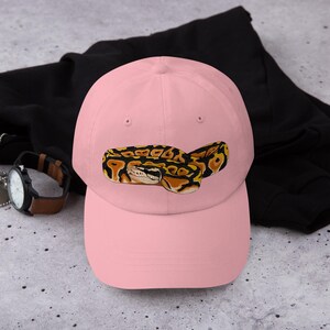 May include: A light pink baseball cap featuring an embroidered image of a ball python. The snake is depicted in shades of yellow, brown, and black. The cap is resting on a black garment.