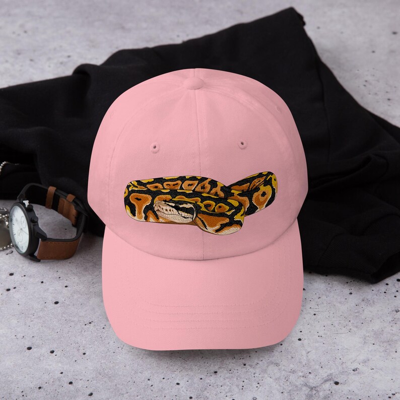 May include: A light pink baseball cap featuring an embroidered image of a ball python. The snake is depicted in shades of yellow, brown, and black. The cap is resting on a black garment.