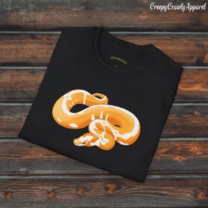 Könnte beinhalten: Schwarzes T-Shirt mit einem weißen und orangefarbenen Schlangenmotiv. Die Schlange ist zusammengerollt und hat ein weißes und orangefarbenes Muster.