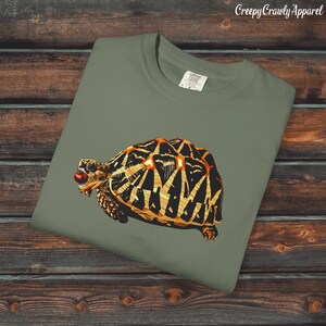 Peut inclure: T-shirt vert olive avec une illustration détaillée d'une tortue. La tortue a une carapace à motifs noirs et dorés et une baie rouge dans la bouche. Le texte "Creepy Crawly Apparel" est dans le coin supérieur droit.