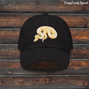 Może przedstawiać: Czarna czapka z daszkiem z żółto-białym wzorem węża. Czapka ma zakrzywiony daszek i strukturalny krój. Tekst "Creepy Crawly Apparel" jest wydrukowany w prawym górnym rogu. Modny dodatek.