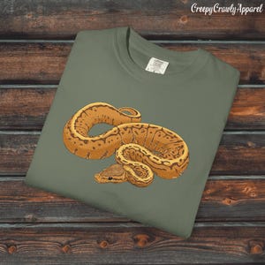 Può includere: T-shirt verde salvia con un'illustrazione dettagliata di un pitone reale marrone e beige. Il serpente è arrotolato e presenta un motivo. Il testo "Creepy Crawly Apparel" si trova nell'angolo in alto a destra.
