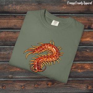 May include: Sage green t-shirt with a detailed illustration of a centipede in red and orange hues. The shirt has a crew neck and a tag at the collar. The text "Creepy Crawly Apparel" is printed at the top.